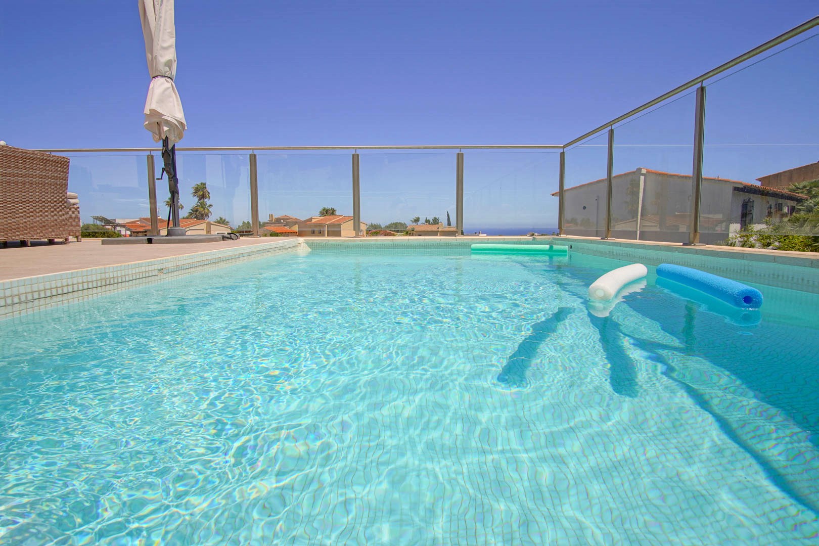 Rectangular swimming pool featuring light blue mosaic tiling and a safety glass balustrade. The sun-drenched terrace includes outdoor furniture and views towards the Mediterranean.