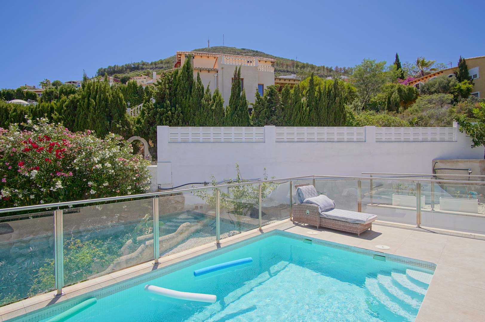 Sun-drenched terrace featuring a rectangular pool with glass safety railings, a woven sun lounger, and a backdrop of mature Mediterranean trees.