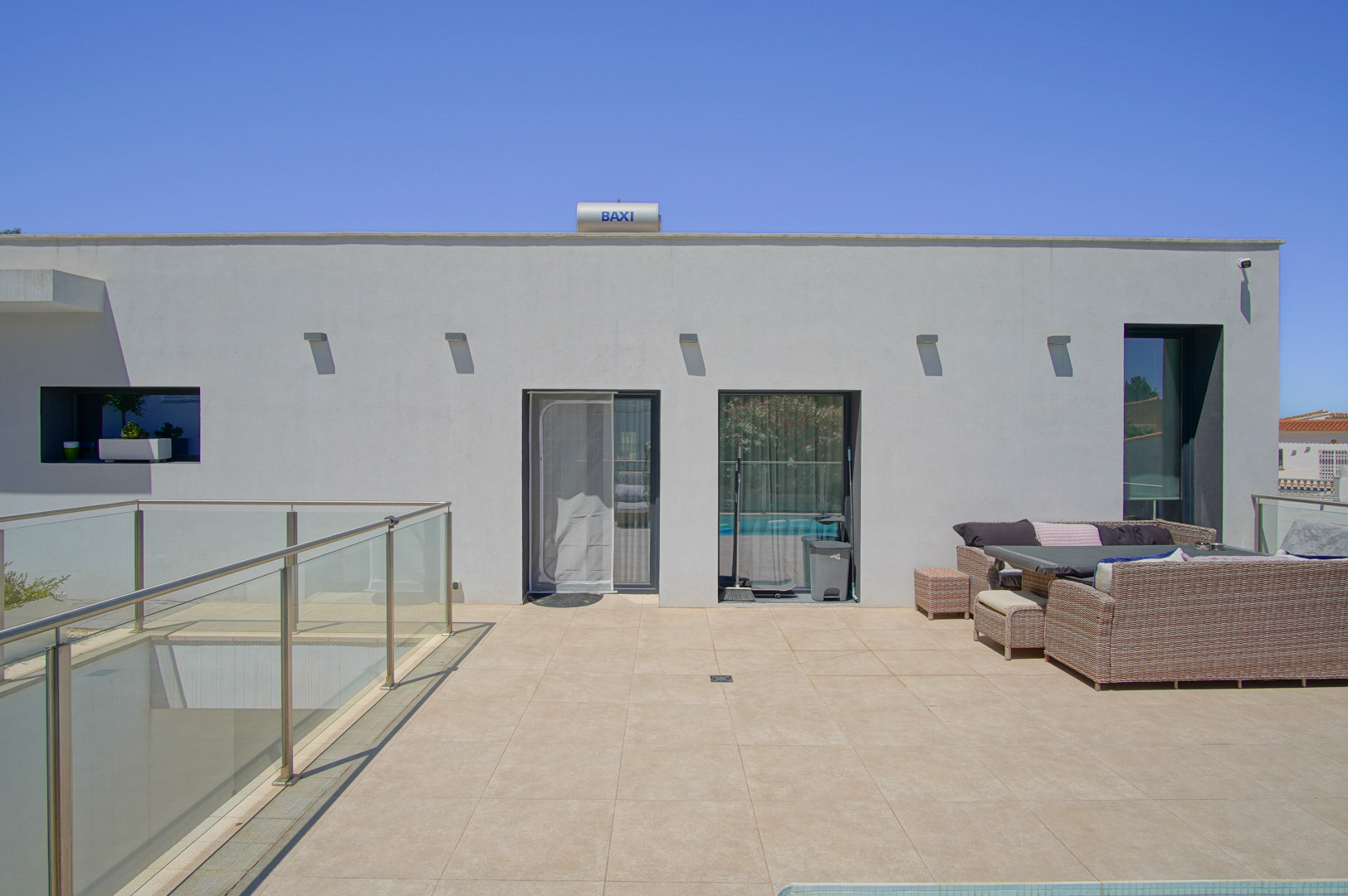 Upper terrace featuring sand-toned ceramic tiling, synthetic rattan outdoor furniture, and glass railings. A BAXI solar water heater is visible on the minimalist white rooftop.