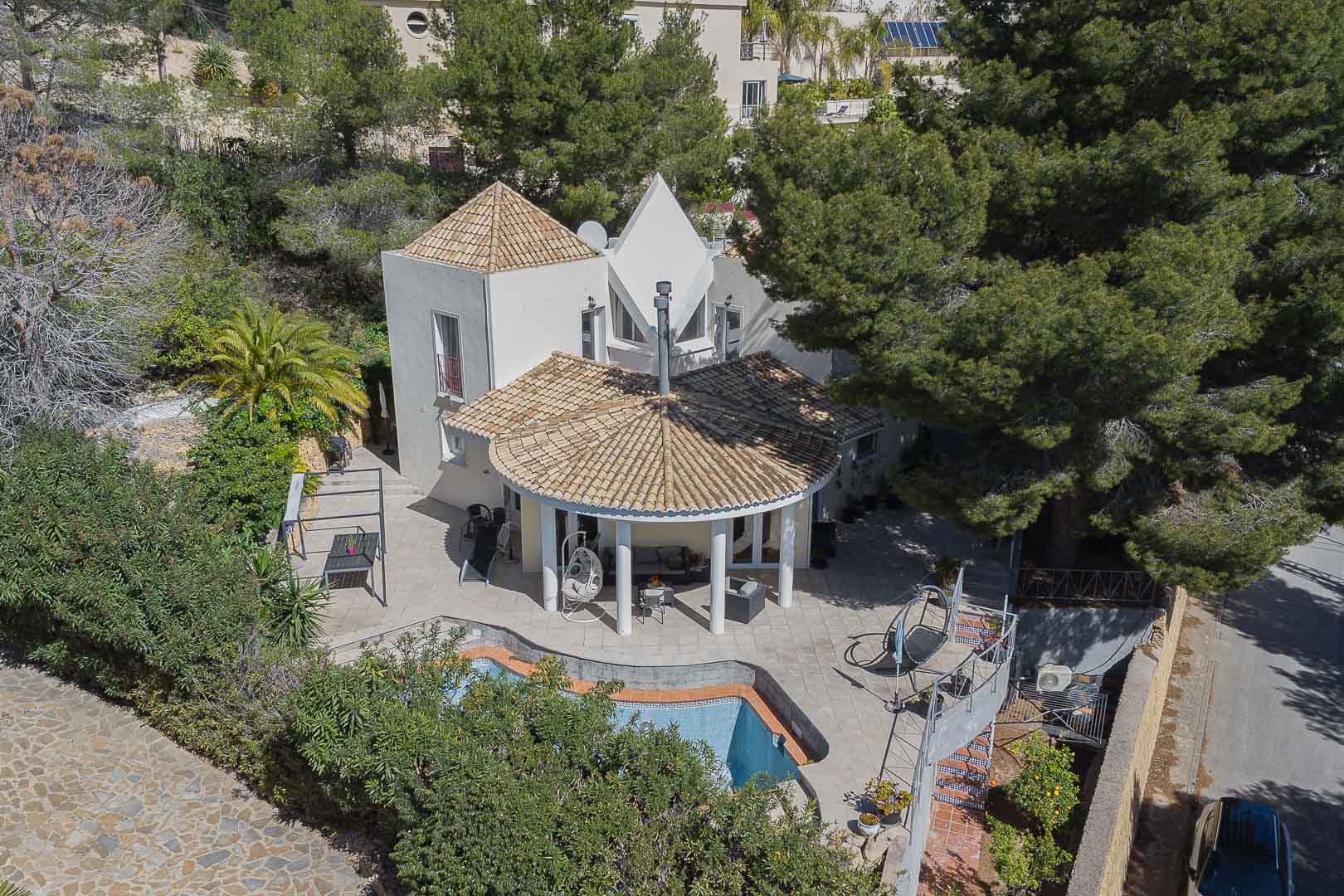 Aerial view of the villa featuring a circular porch with white columns, a private pool, and stone-paved terraces nestled among mature pine trees and gardens.