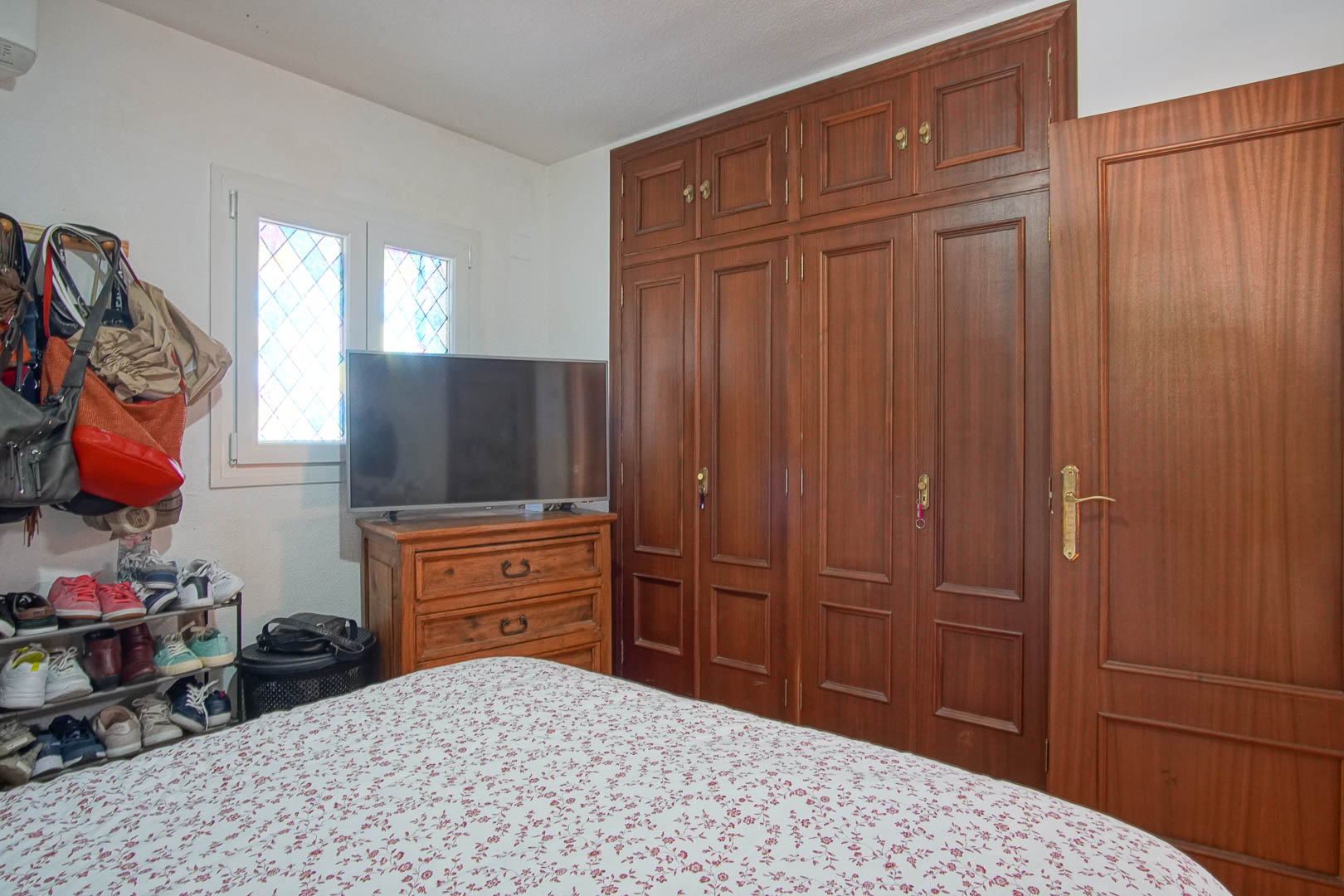 Bedroom featuring a large floor-to-ceiling wooden wardrobe, matching dresser with TV, and a window with traditional leaded glass detailing.
