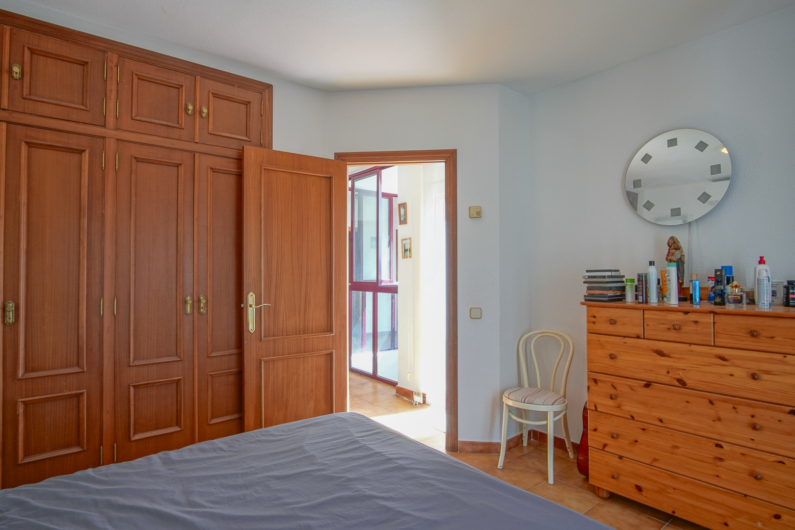 Bedroom featuring floor-to-ceiling built-in wooden wardrobes, a matching chest of drawers, and tiled flooring. The space opens onto a bright hallway through a classic wooden door.