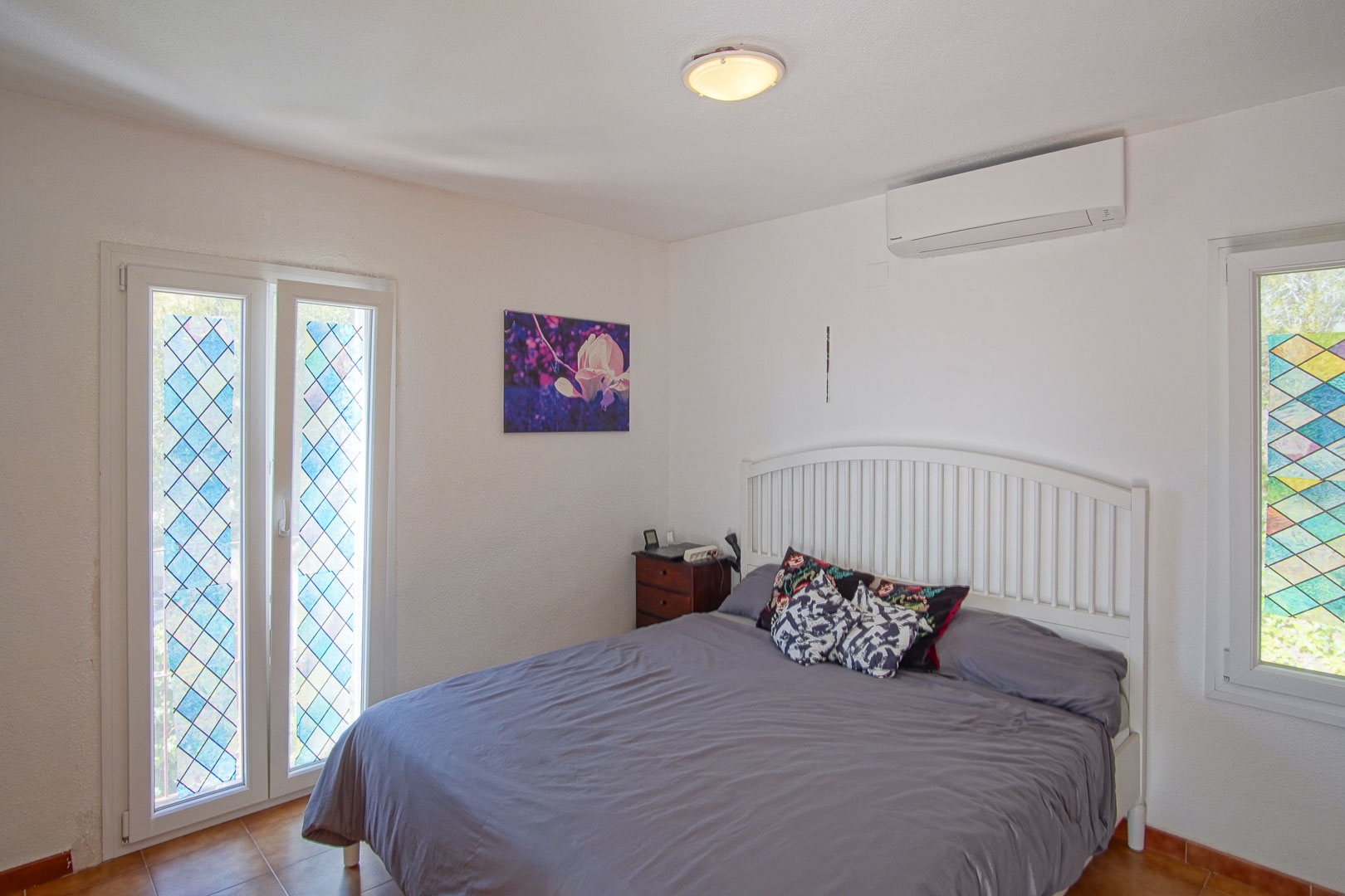Bright bedroom featuring split air conditioning, a white slatted headboard, and windows with decorative diamond-patterned stained glass.