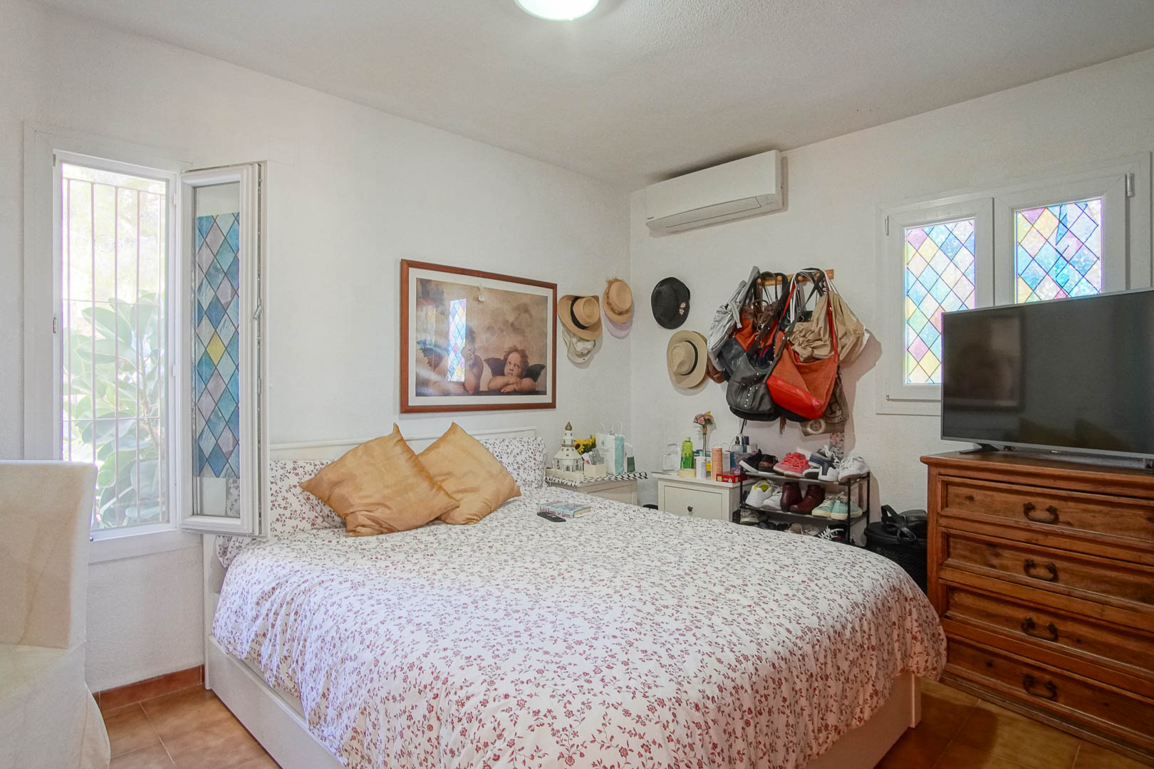 Bright bedroom featuring terracotta tile flooring, split air conditioning, and windows with decorative stained glass. It includes a classic wooden dresser and natural light.