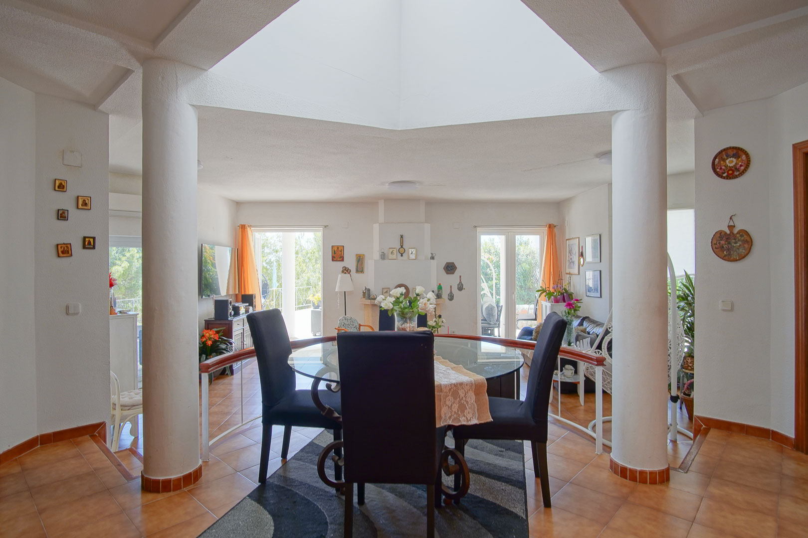 Bright dining area featuring a central skylight and white columns, with terracotta tiling and a view toward the living room and fireplace.