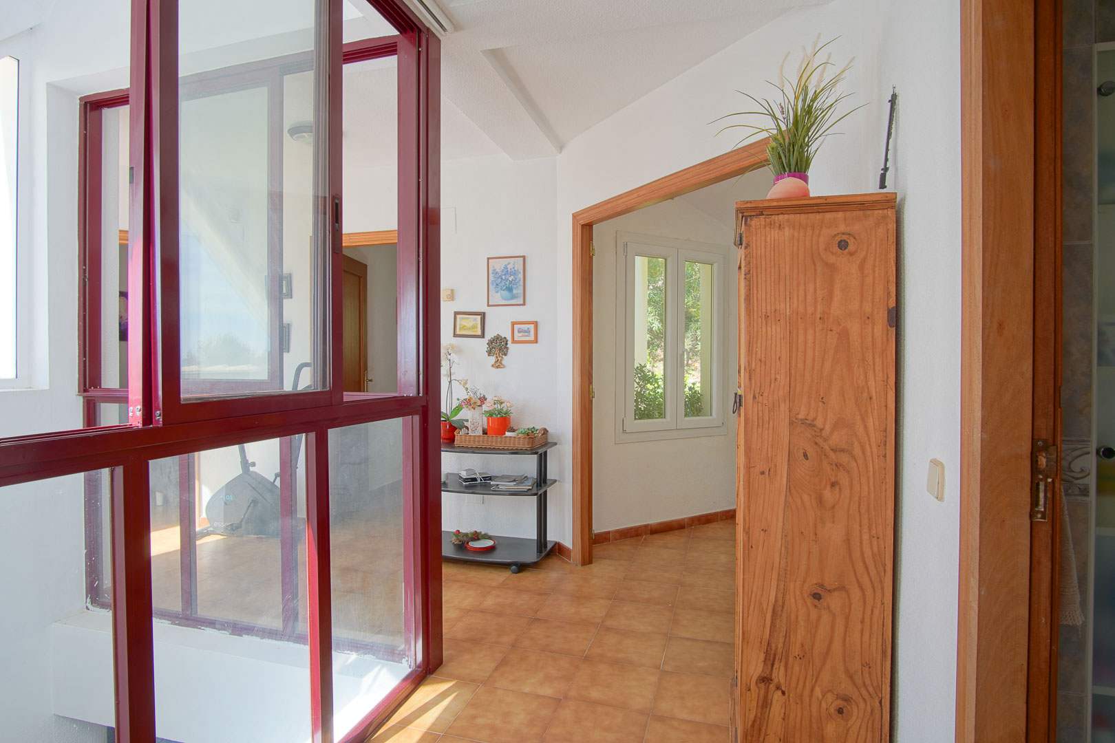 Bright landing featuring terracotta tiles, wooden door frames, and a glass enclosure with burgundy frames overlooking an indoor exercise area.