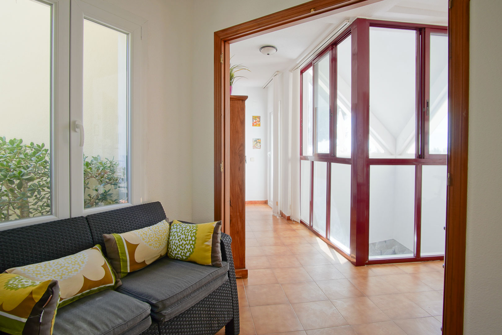 Bright seating area featuring a wicker sofa, terracotta tile flooring, and a glass-enclosed sunroom with distinctive burgundy frames.
