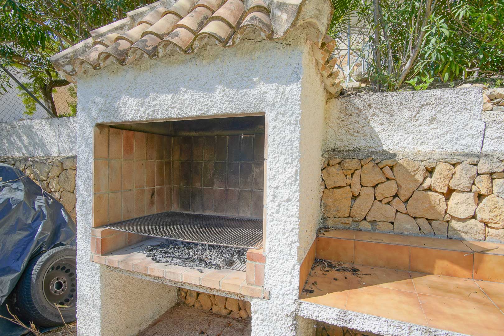 Built-in outdoor barbecue featuring a terracotta tile roof and white stucco finish, situated against a natural stone wall with an adjacent tiled preparation area.