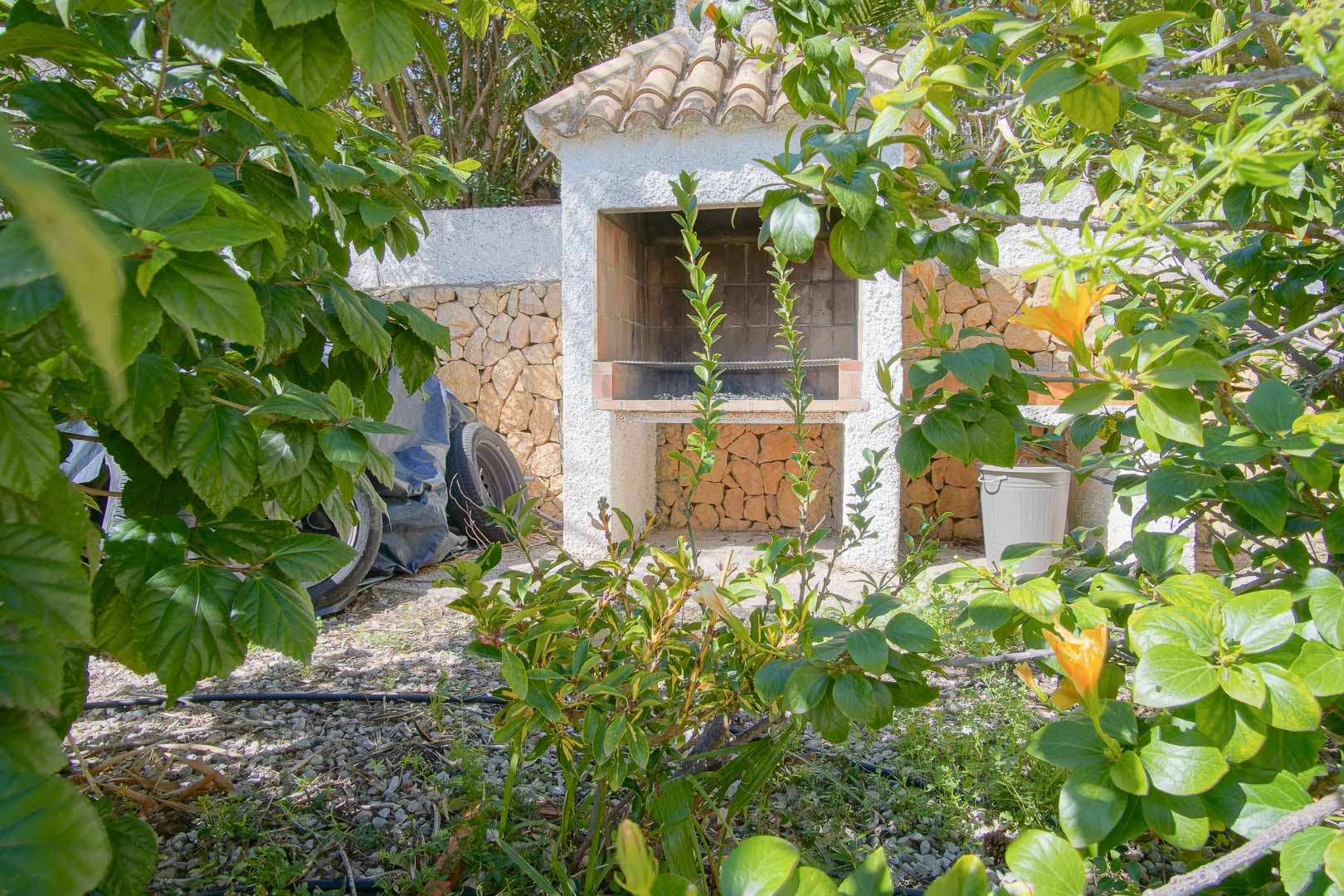 Built-in outdoor barbecue station featuring a traditional tiled roof, set against a natural stone wall and surrounded by lush garden greenery.