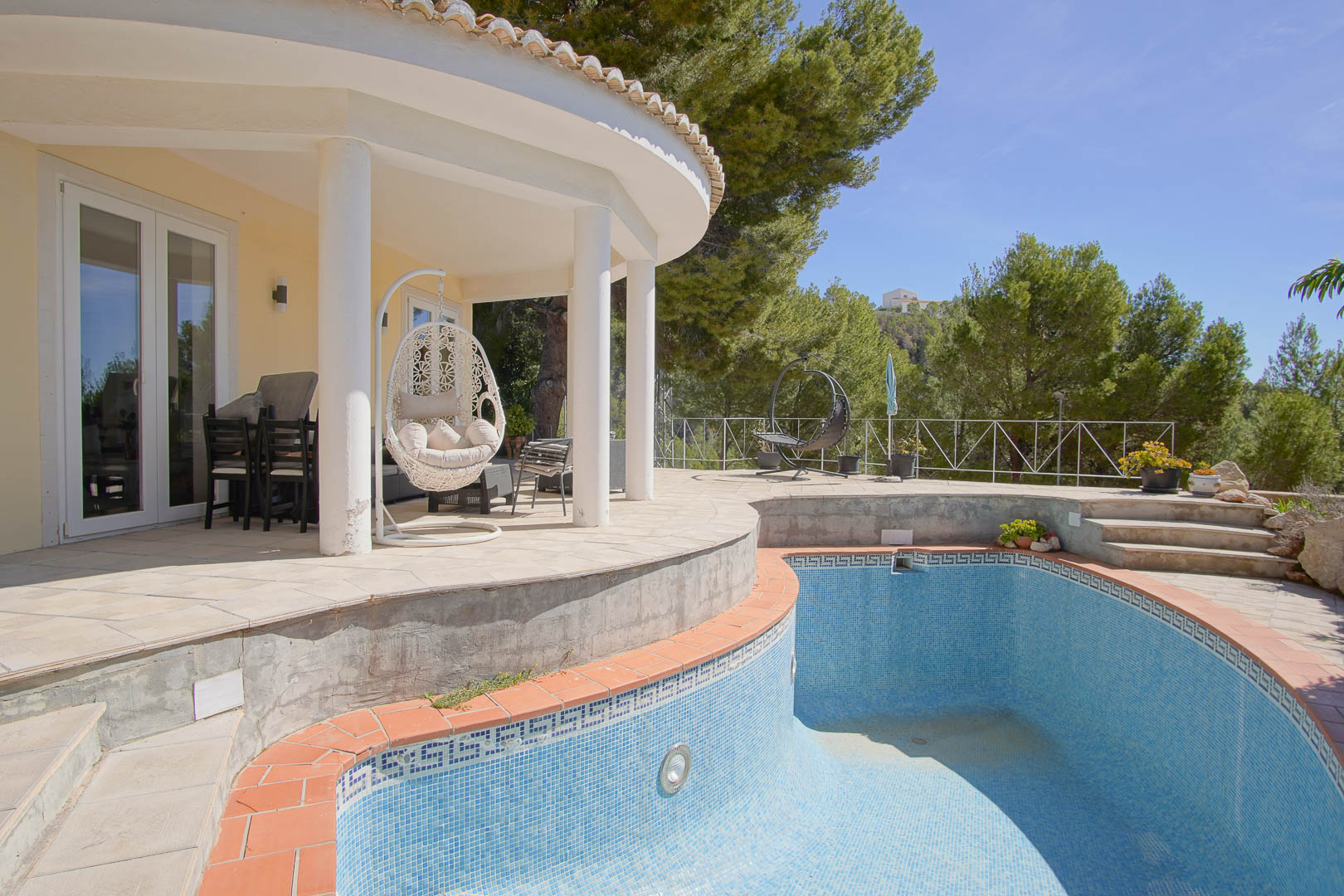Curved covered porch with white columns and a hanging chair, overlooking a mosaic-tiled pool area bordered by mature pine trees and natural scenery.