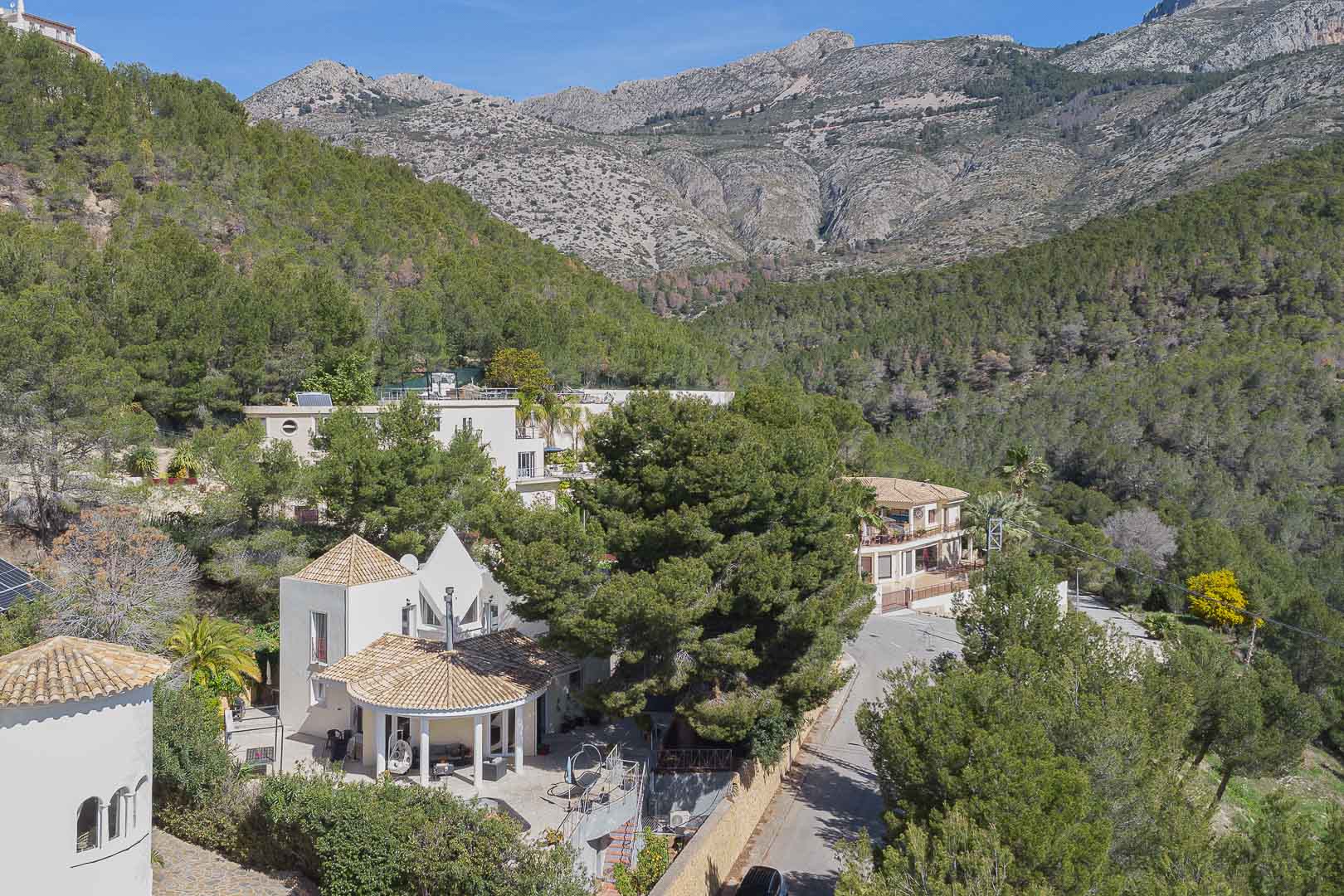 Elevated view of the villa featuring Mediterranean architecture with tiled roofs, nestled within a lush pine forest against a dramatic mountain backdrop.