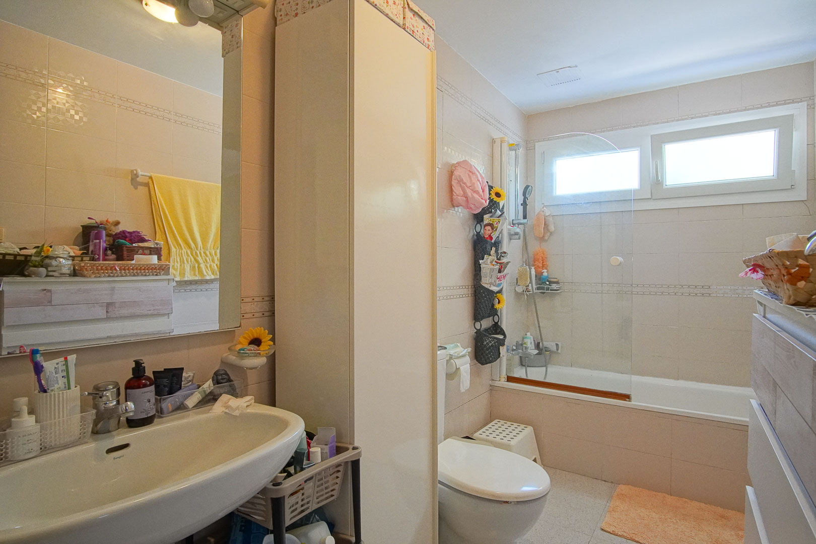 Full bathroom featuring a bathtub with glass screen, white sink, and tall storage cabinet. Cream wall tiles and a window provide natural light and ventilation.