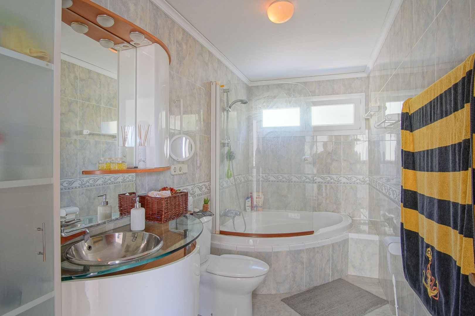 Full bathroom featuring a corner bathtub with glass screen and a modern stainless steel vessel sink on a glass vanity. Tiled walls with decorative borders and natural light.