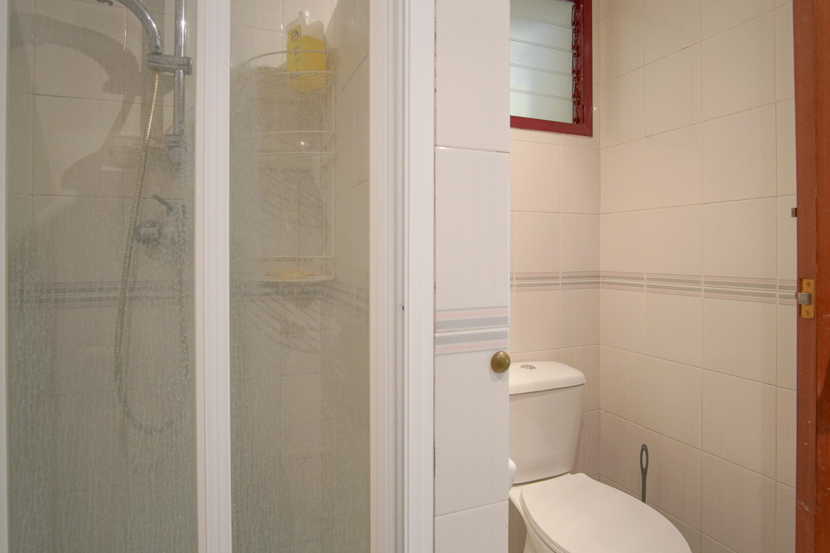 Functional bathroom featuring white tiled walls with decorative borders, a glass-enclosed shower, and a high-set ventilation window with a red frame.