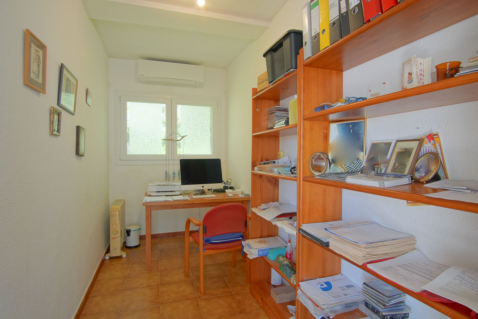 Functional home office featuring a wooden desk, extensive shelving, terracotta tile flooring, and wall-mounted air conditioning for a comfortable workspace.