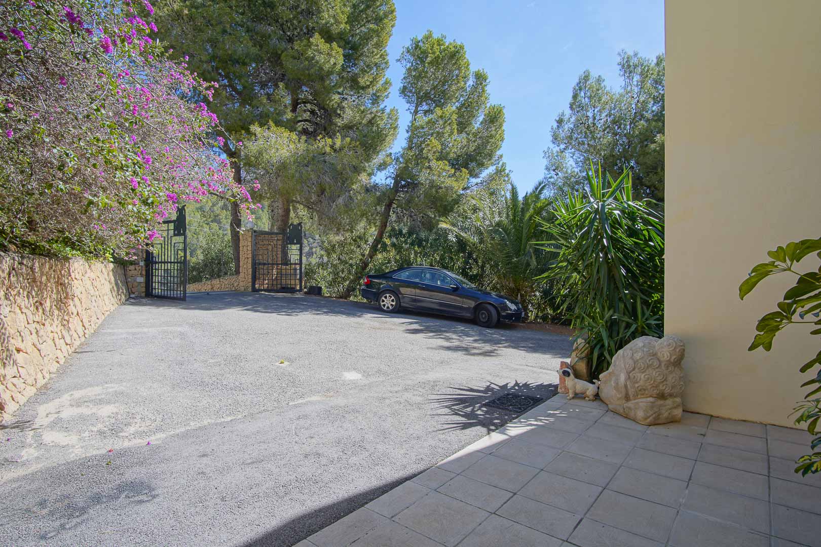 Gated entrance with a paved driveway providing ample outdoor parking, framed by natural stone walls and mature Mediterranean trees.
