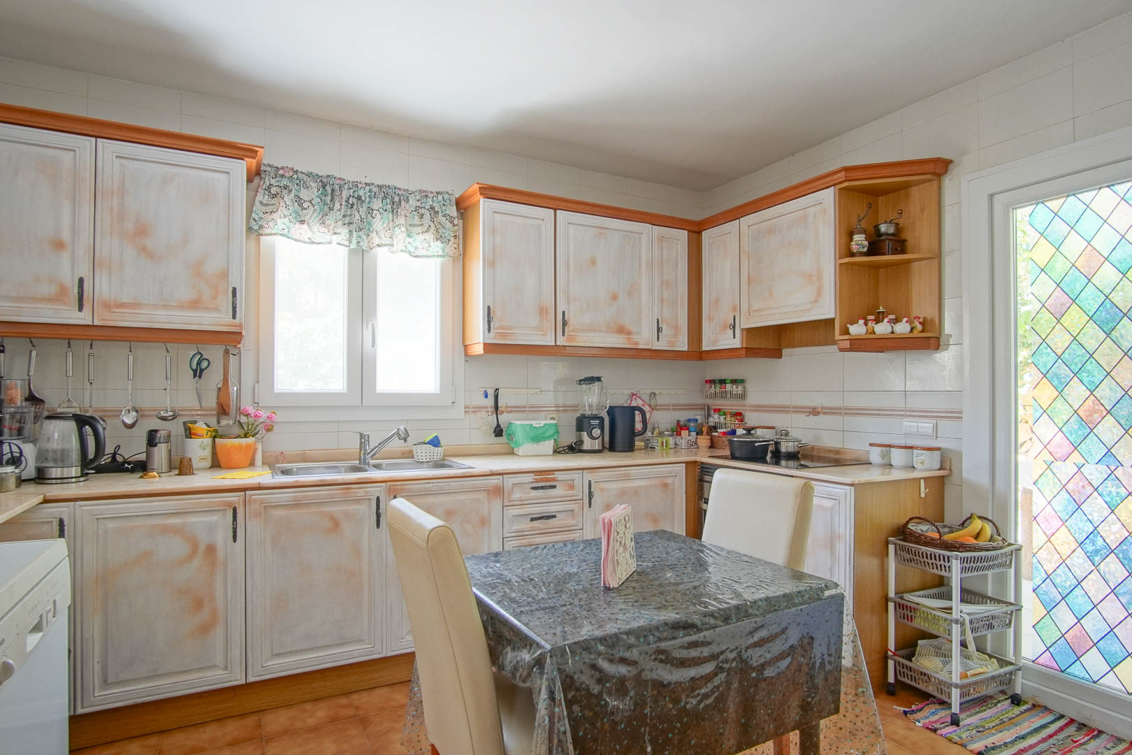 Independent rustic kitchen featuring distressed-finish cabinetry, an L-shaped layout, and a dining nook. It offers natural light and access through a decorative stained-glass door.
