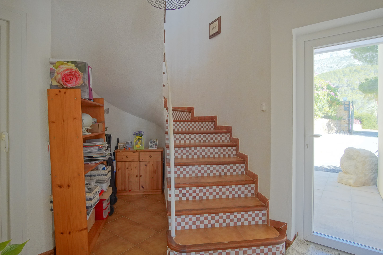 Interior staircase featuring terracotta tiles and checkered risers, complemented by pine wood furniture and a glass door leading to the garden.