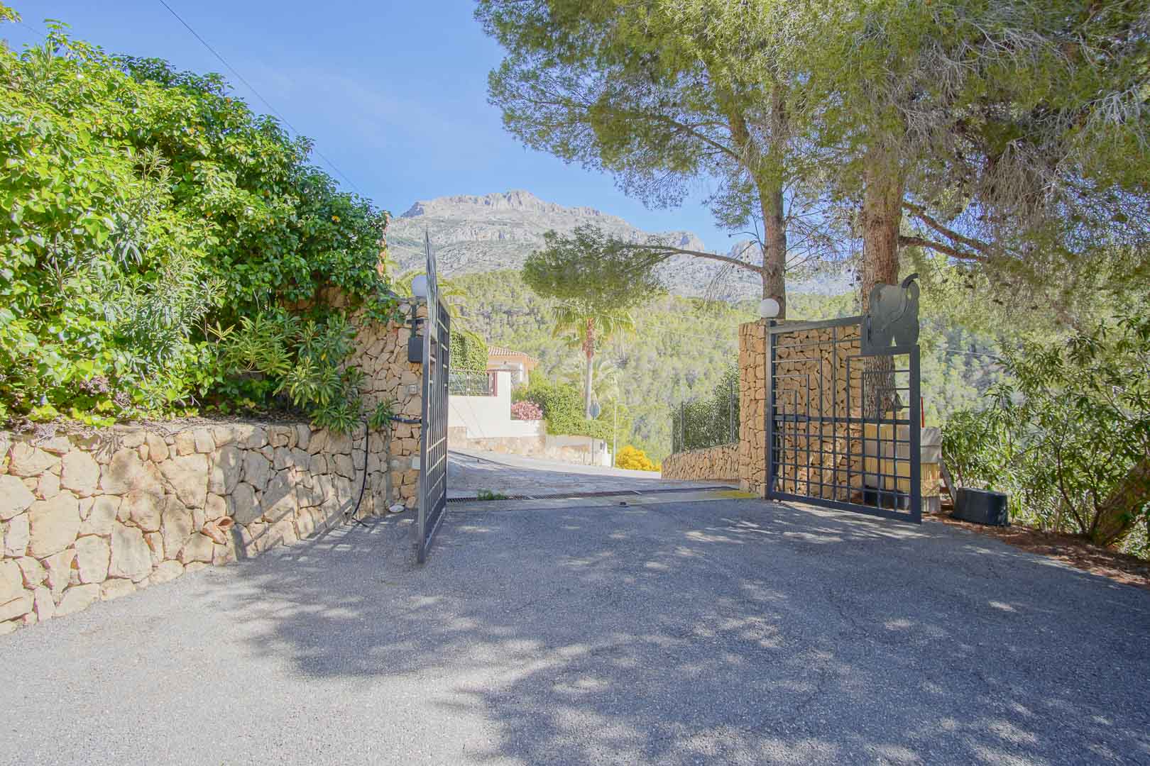 Main entrance featuring a wrought iron gate and natural stone walls, opening onto a paved driveway with views of the surrounding mountains and pine trees.