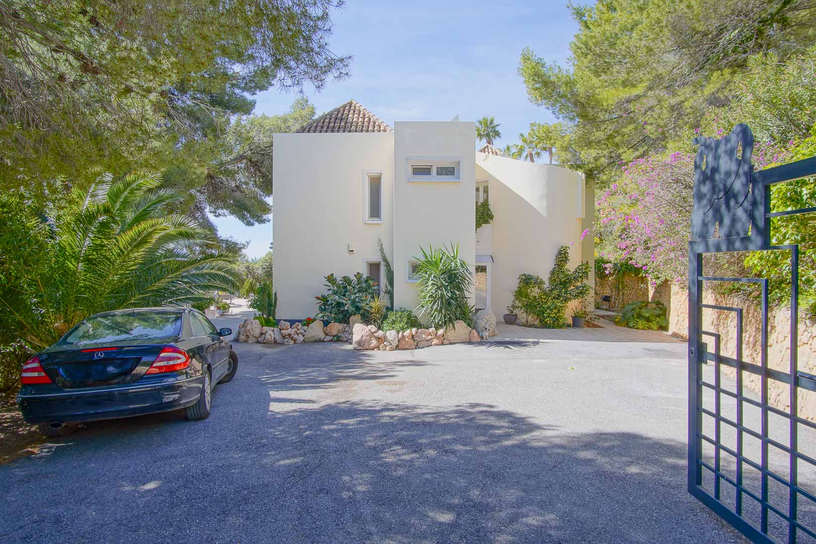 Main facade of the villa featuring Mediterranean architecture, a spacious paved parking area, and a gated entrance surrounded by mature pine trees and lush greenery.
