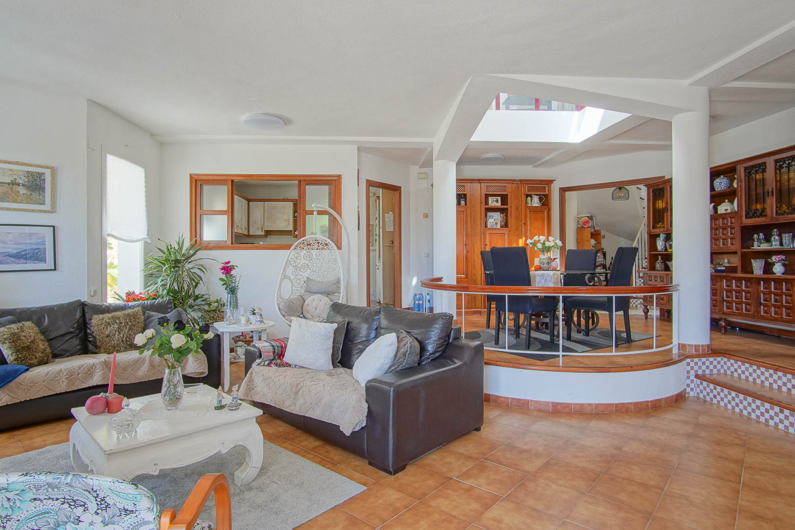 Open-plan living area featuring terracotta tiles, leather sofas, and an elevated dining space with a circular railing and natural light from a central skylight.