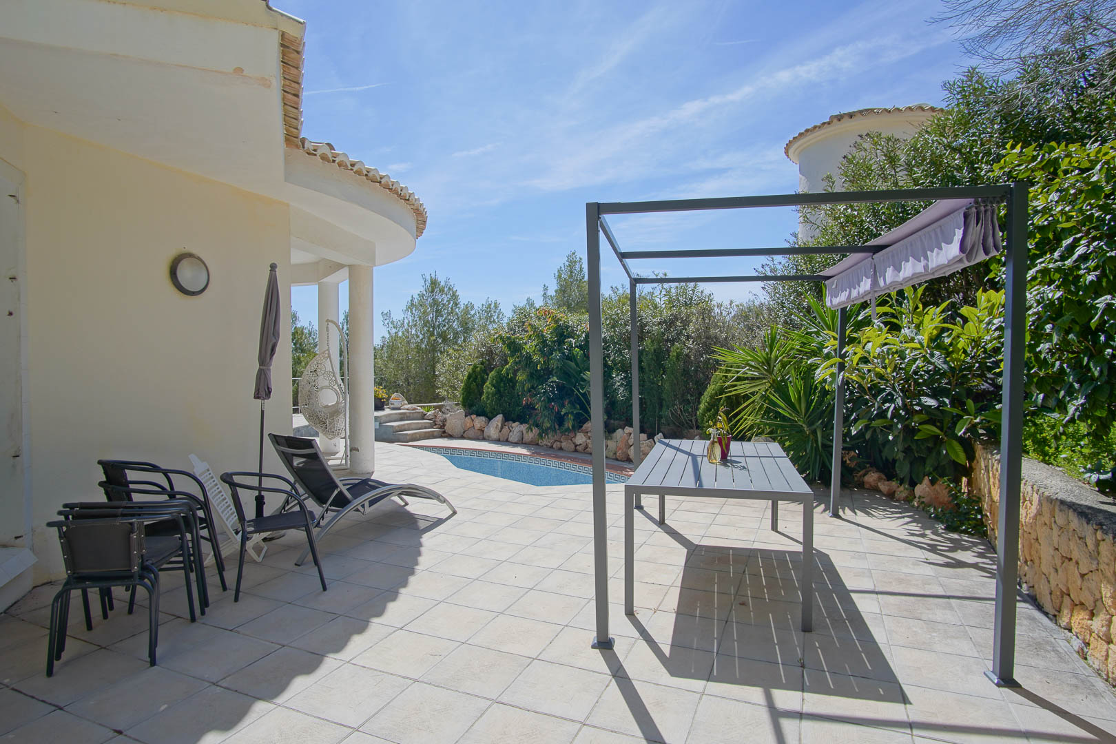 Outdoor terrace featuring tiled flooring, a metal pergola with a dining set, and a private swimming pool bordered by mature Mediterranean gardens.