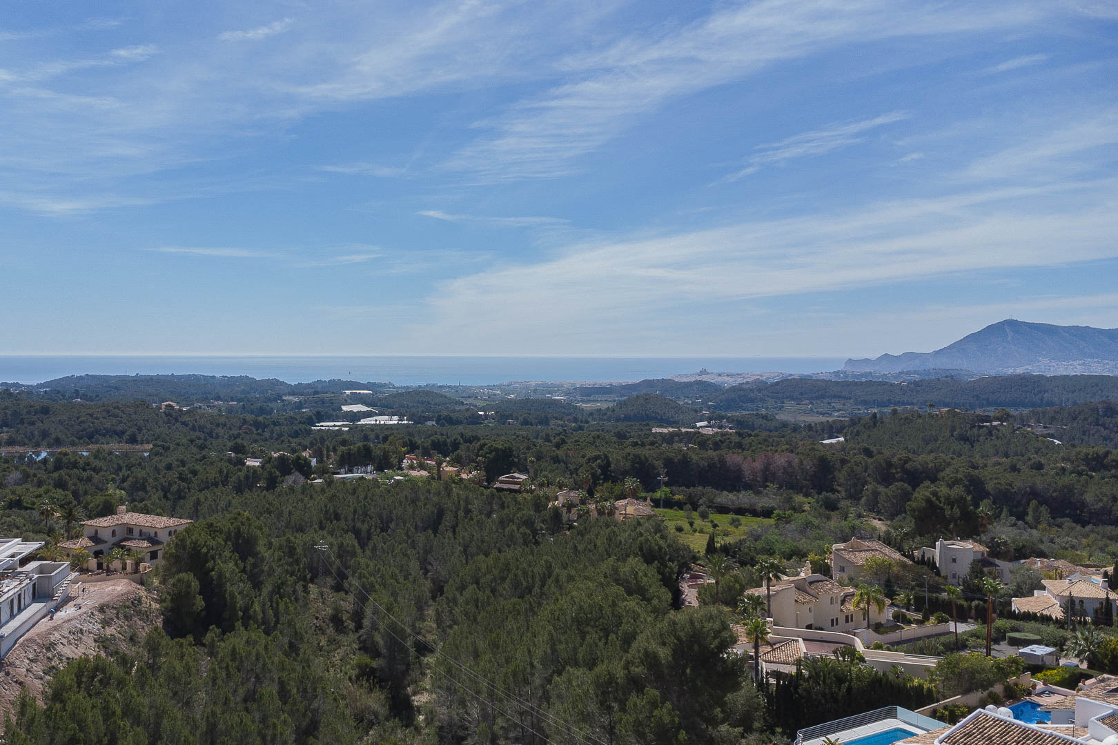 Panoramic view from the upper terrace overlooking a lush pine forest, the Mediterranean coastline on the horizon, and mountain silhouettes under a clear sky.