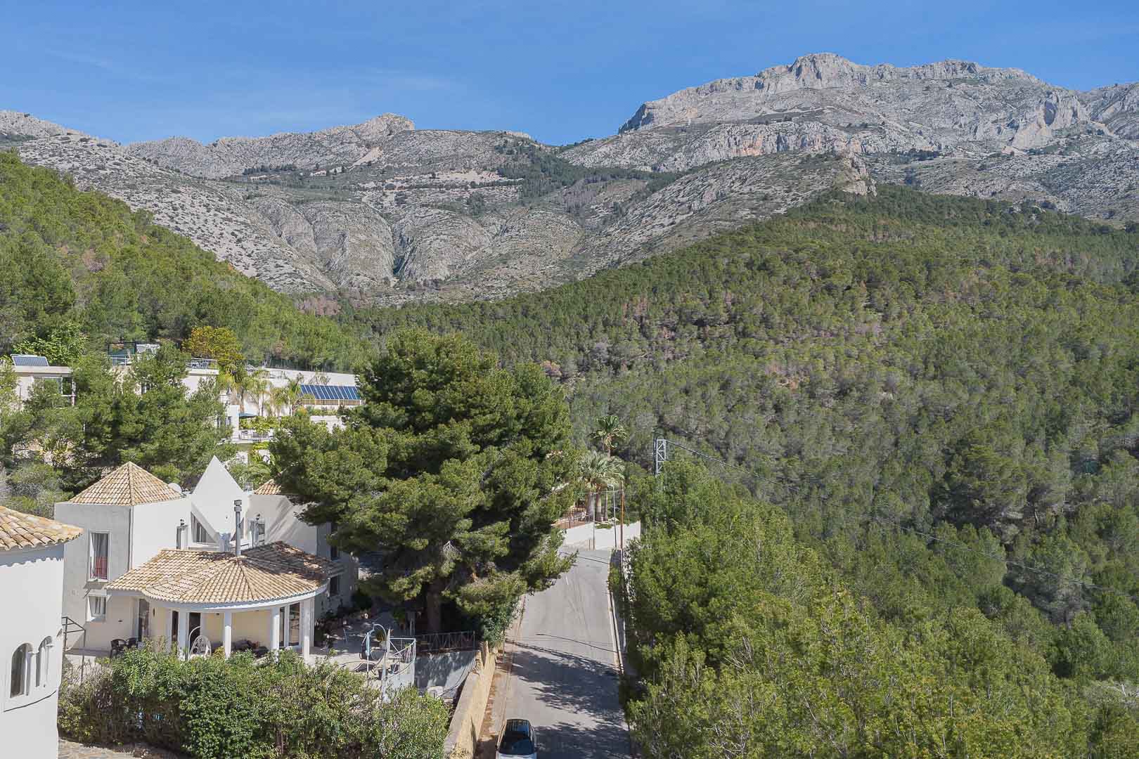 Panoramic view of the villa nestled in nature, showcasing its Mediterranean architecture against a dense pine forest and the majestic mountain range.