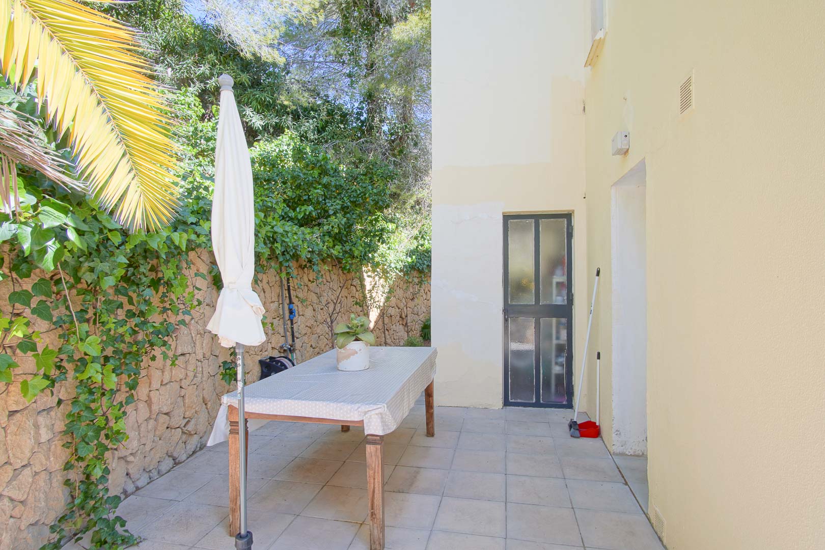 Paved outdoor patio featuring a natural stone wall with ivy, a wooden dining table, and a parasol, situated adjacent to the villa's side entrance.