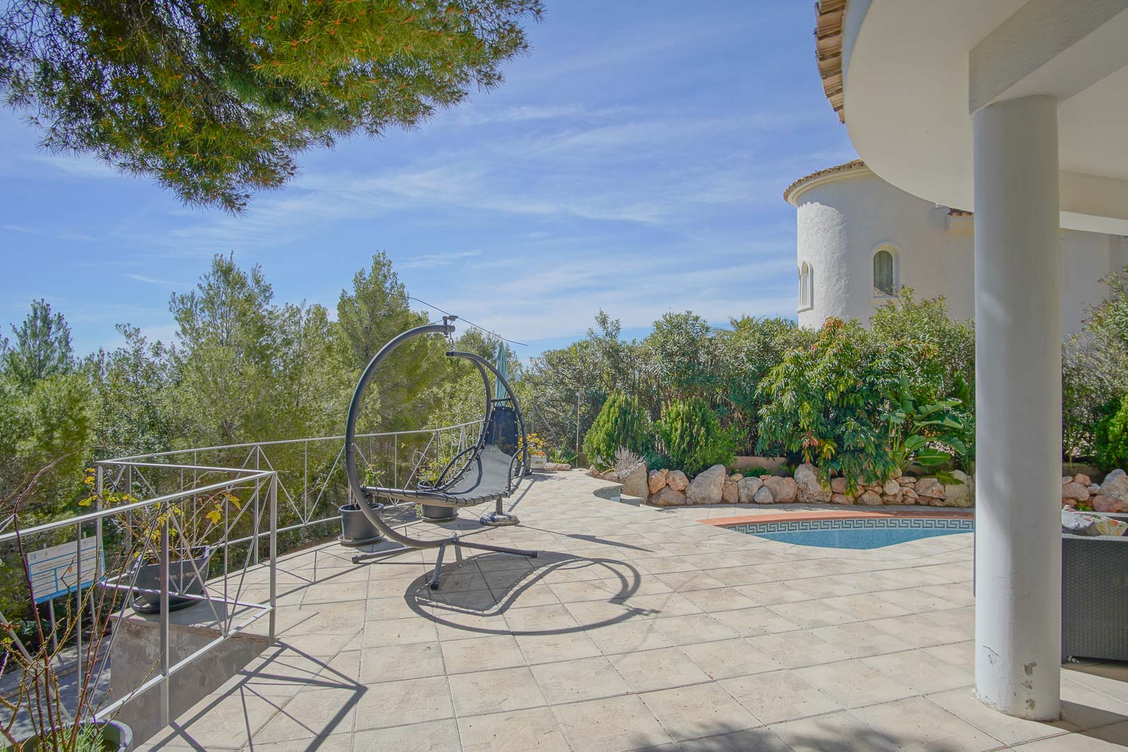 Paved outdoor terrace featuring a modern hanging chair, direct pool access with mosaic detailing, and views of the lush Mediterranean landscape.