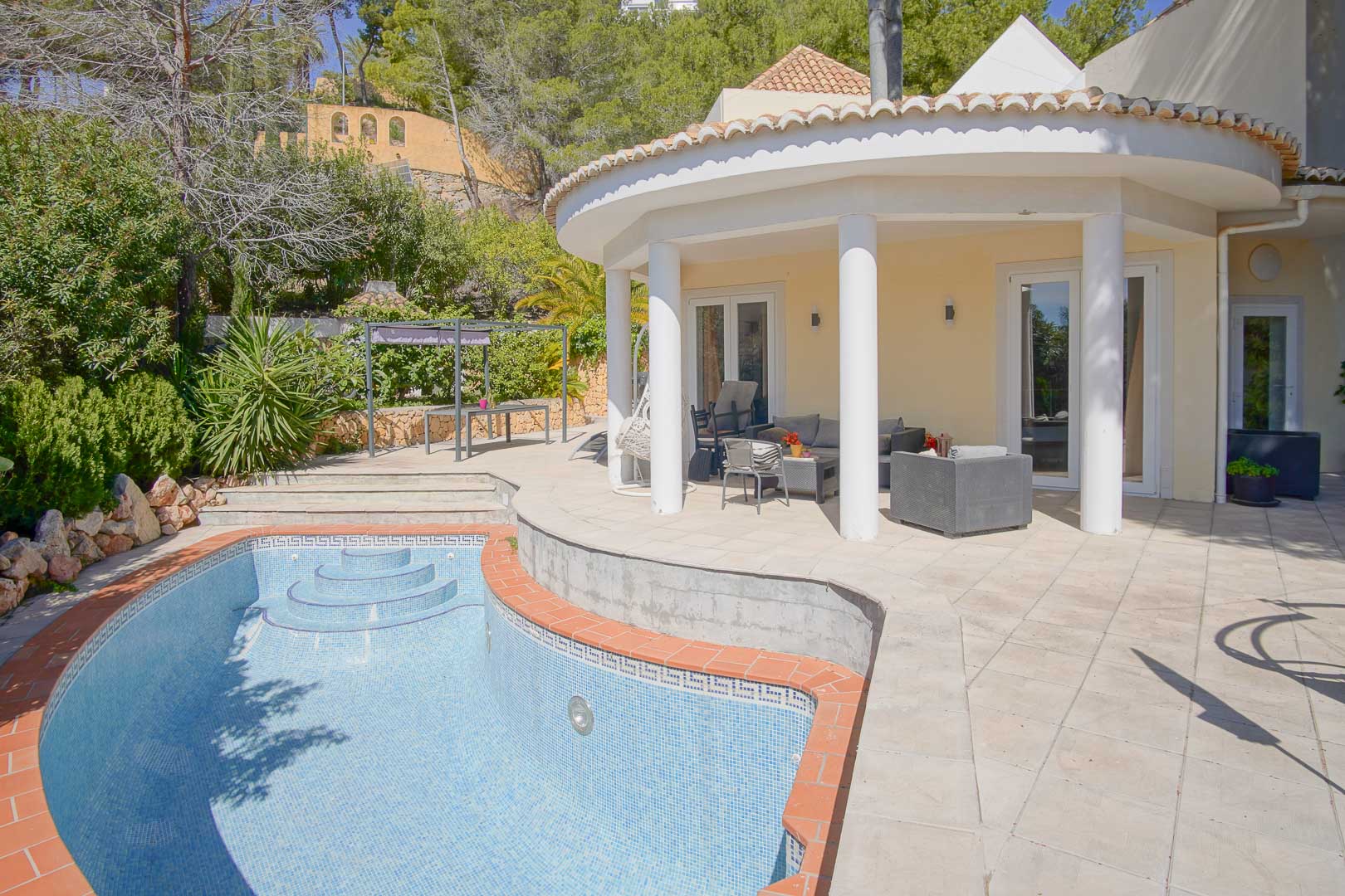 Semi-circular covered porch with white columns overlooking a private mosaic-tiled pool and sun terrace, set within a lush Mediterranean garden.