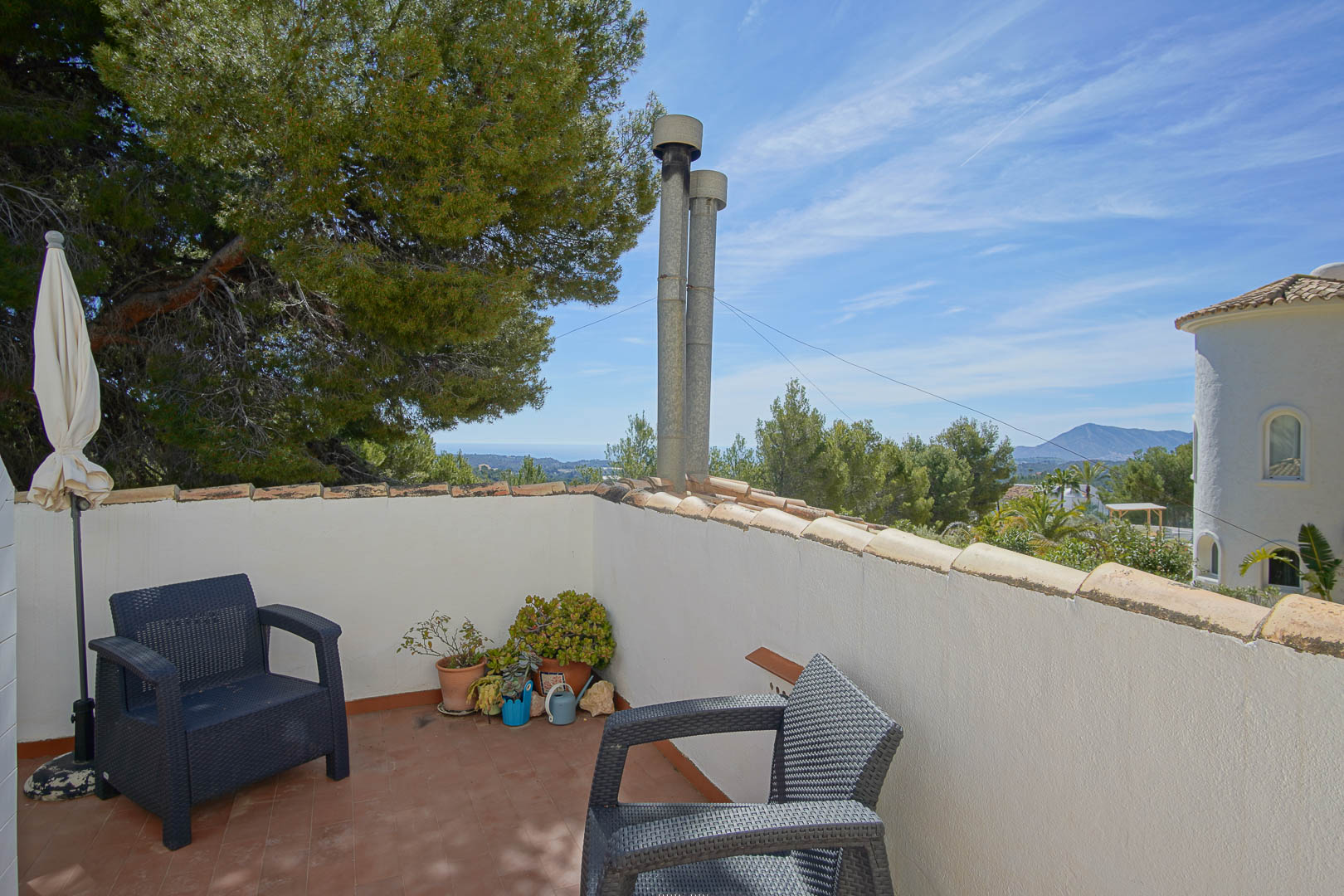 Upper terrace featuring terracotta tiling and wicker-style seating, overlooking the Mediterranean Sea and surrounding pine forests under a clear sky.
