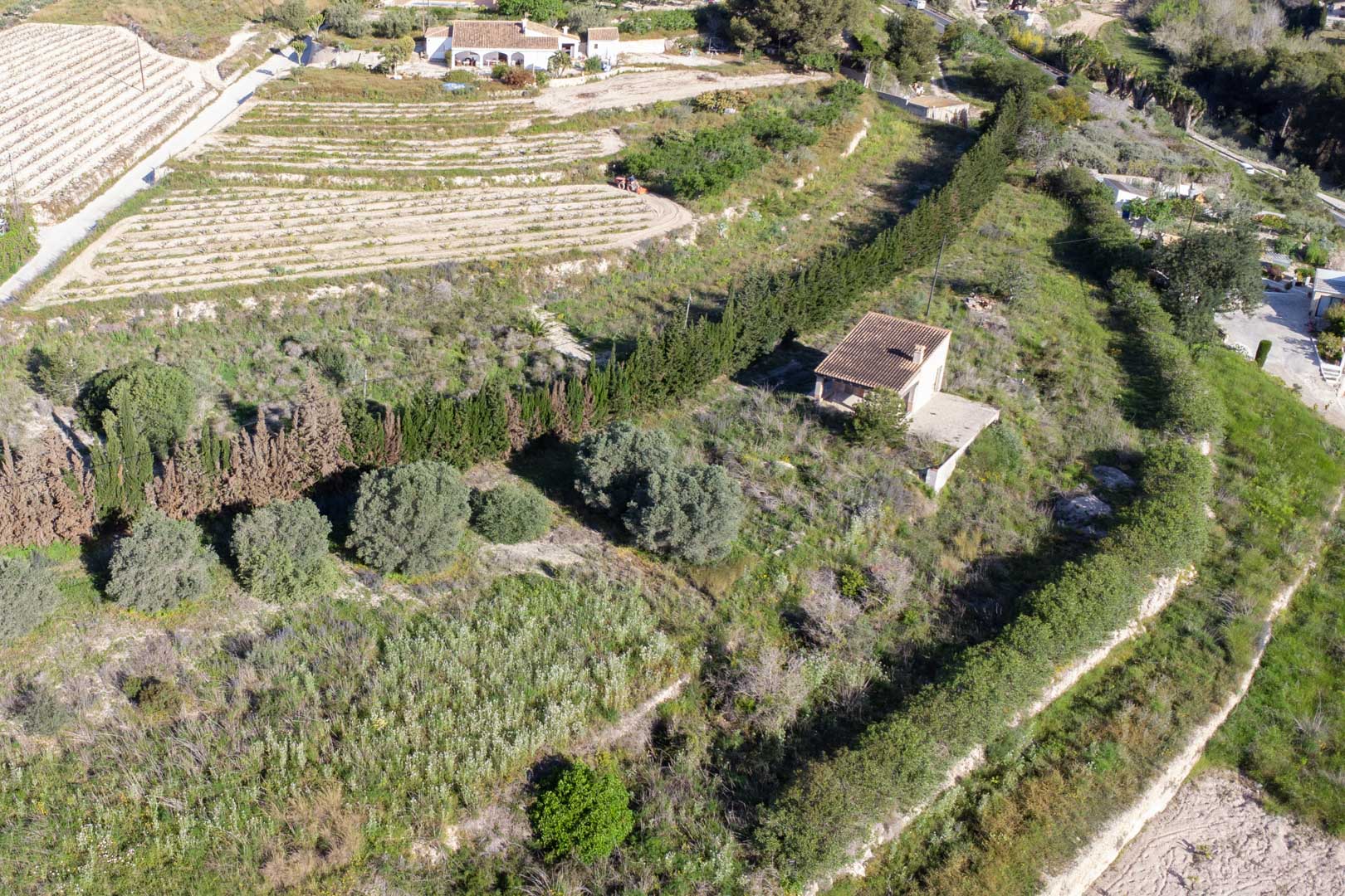 Aerial view of the 10,920 m2 plot in Teulada, featuring terraced land with Mediterranean vegetation and an existing rural outbuilding.