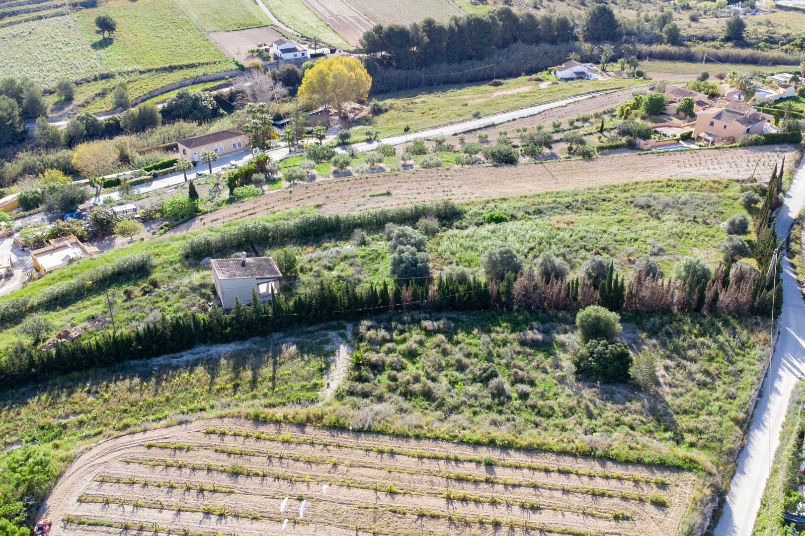 Aerial view of the expansive rustic plot in Teulada, featuring terraced land with Mediterranean vegetation and an existing small outbuilding.