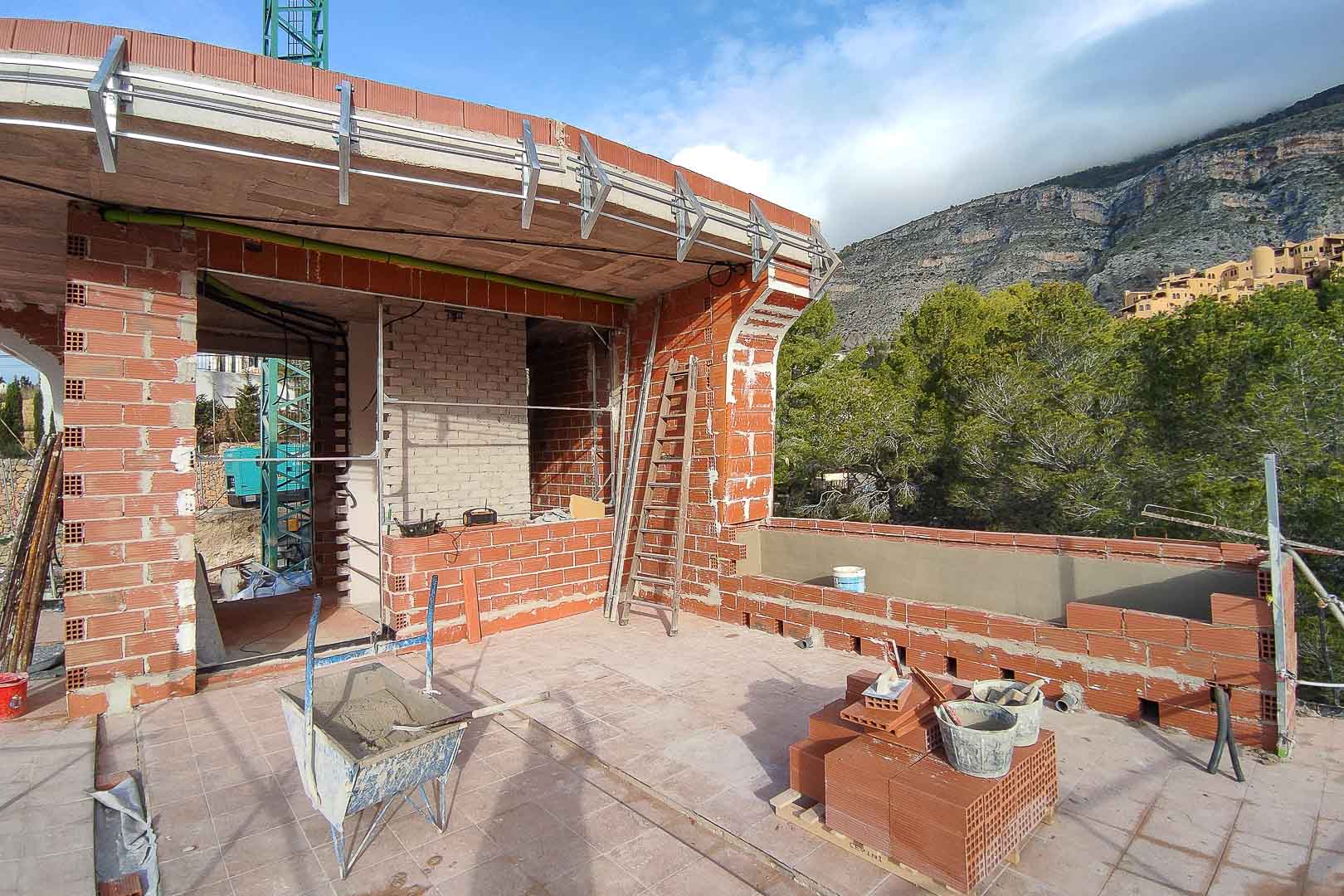 Construction phase of the upper terrace featuring red brick masonry, concrete slabs, and exterior metal framing against a backdrop of Mediterranean pine trees.