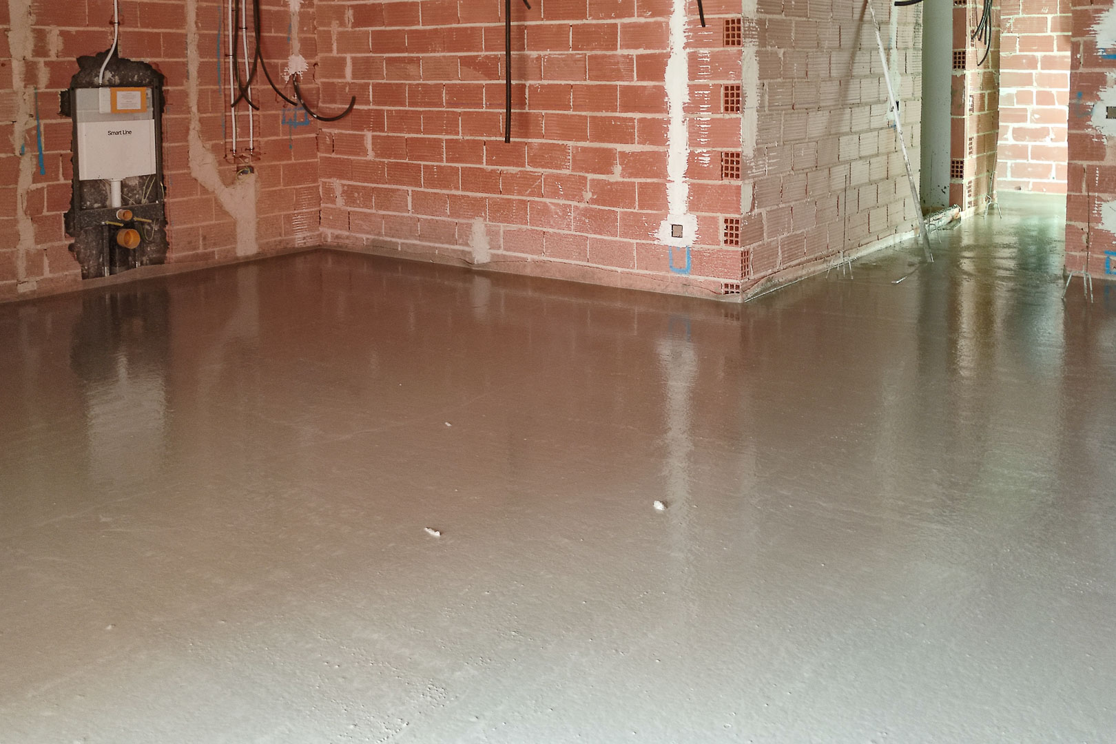 Construction phase showing freshly poured self-leveling floor screed. Visible brickwork and recessed cistern installation for a wall-mounted toilet.