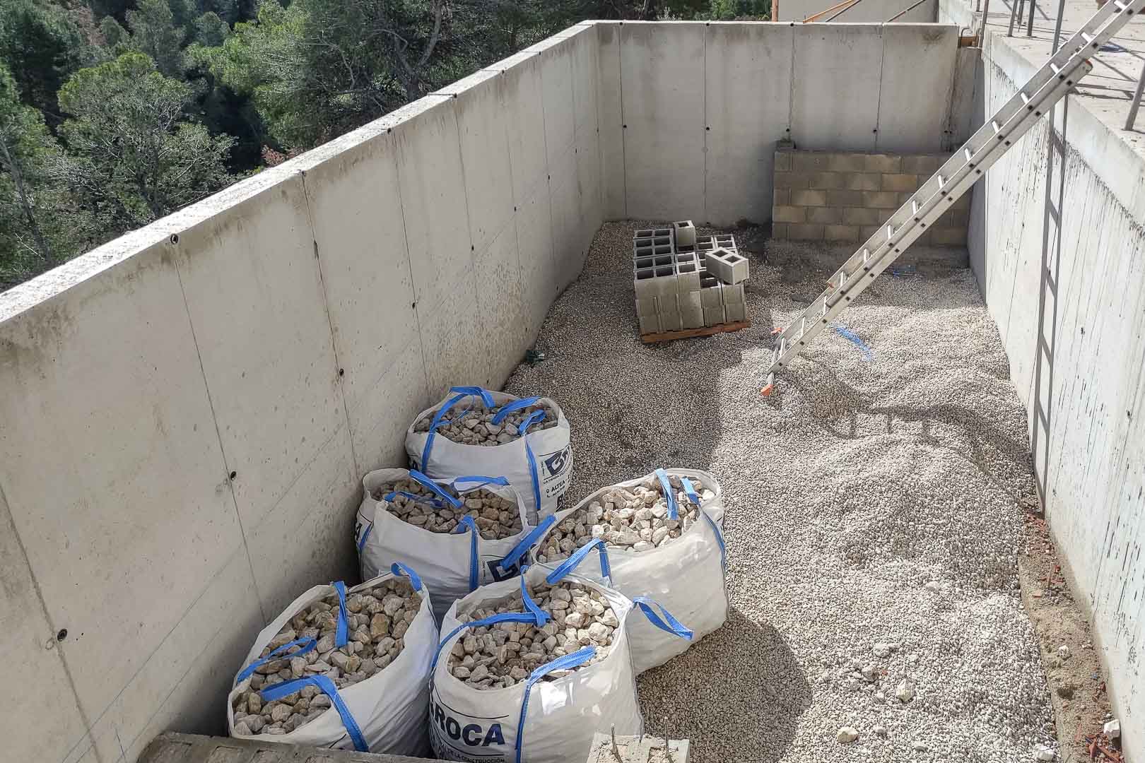 Construction progress showing reinforced concrete walls, foundation gravel, and masonry supplies integrated into the natural hillside landscape of Altea.