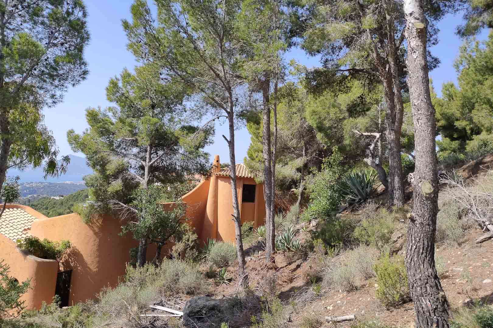Exterior view of the organic terracotta-toned architecture, nestled within a dense pine forest and native Mediterranean vegetation with distant sea views.