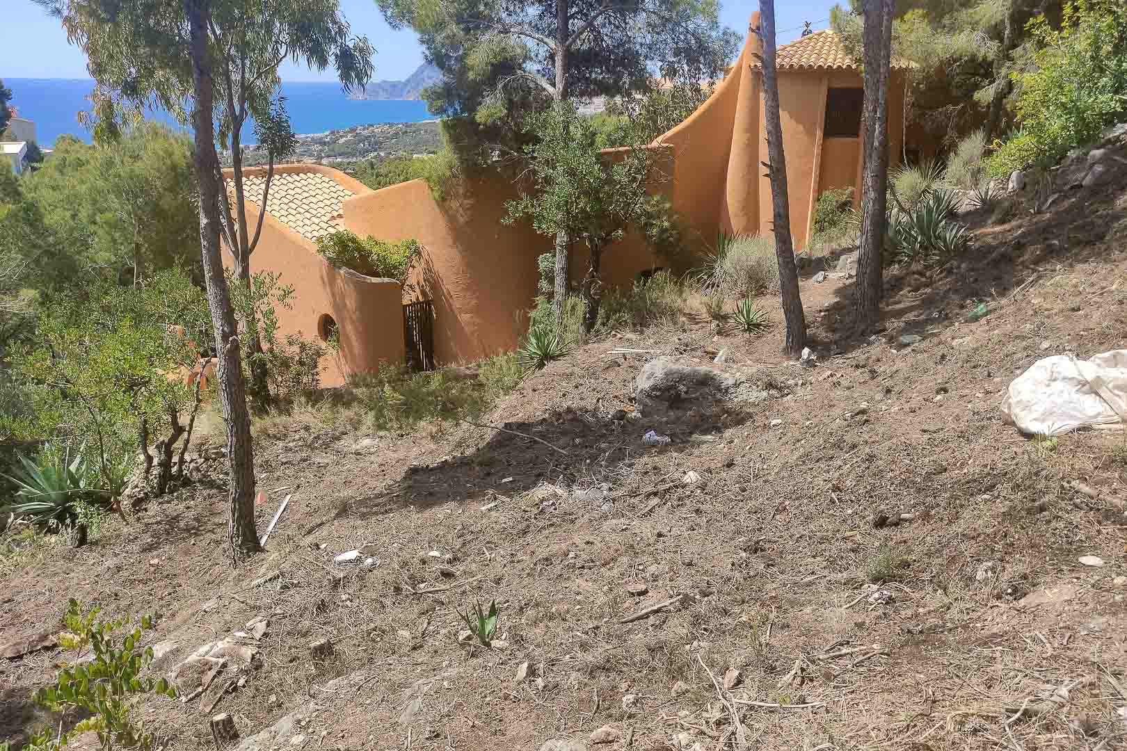 Exterior view of the villa featuring organic architecture and terracotta facades, set on a sloped plot with pine trees and panoramic Mediterranean Sea views.