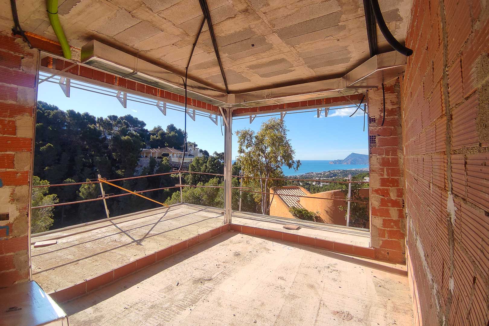 Panoramic sea and mountain views from a room under construction, featuring large corner window frames and exposed HVAC installations.