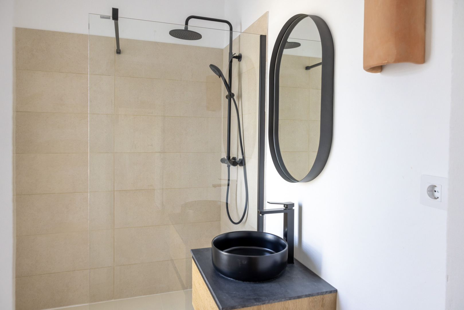 Bathroom detail with beige tiled walk-in shower, black vessel sink on slate countertop, and oval mirror.