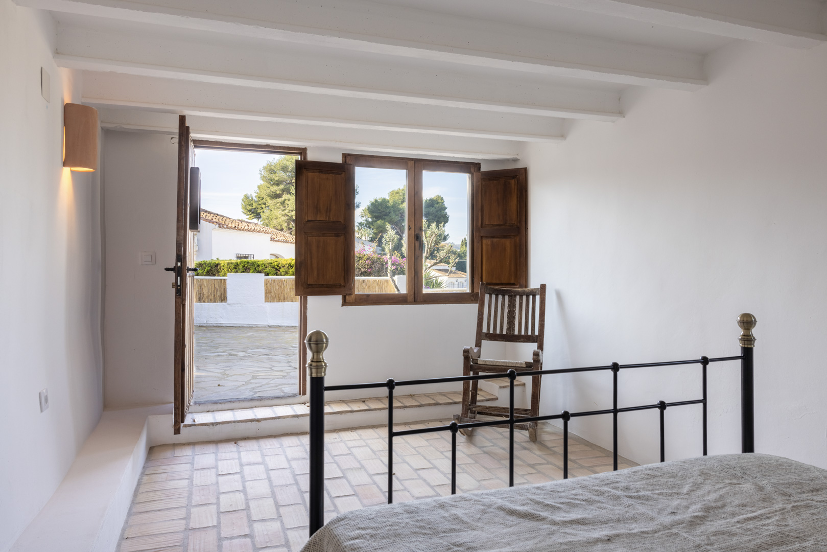 Bright bedroom featuring terracotta flooring, a patio door, and a window with wooden shutters.