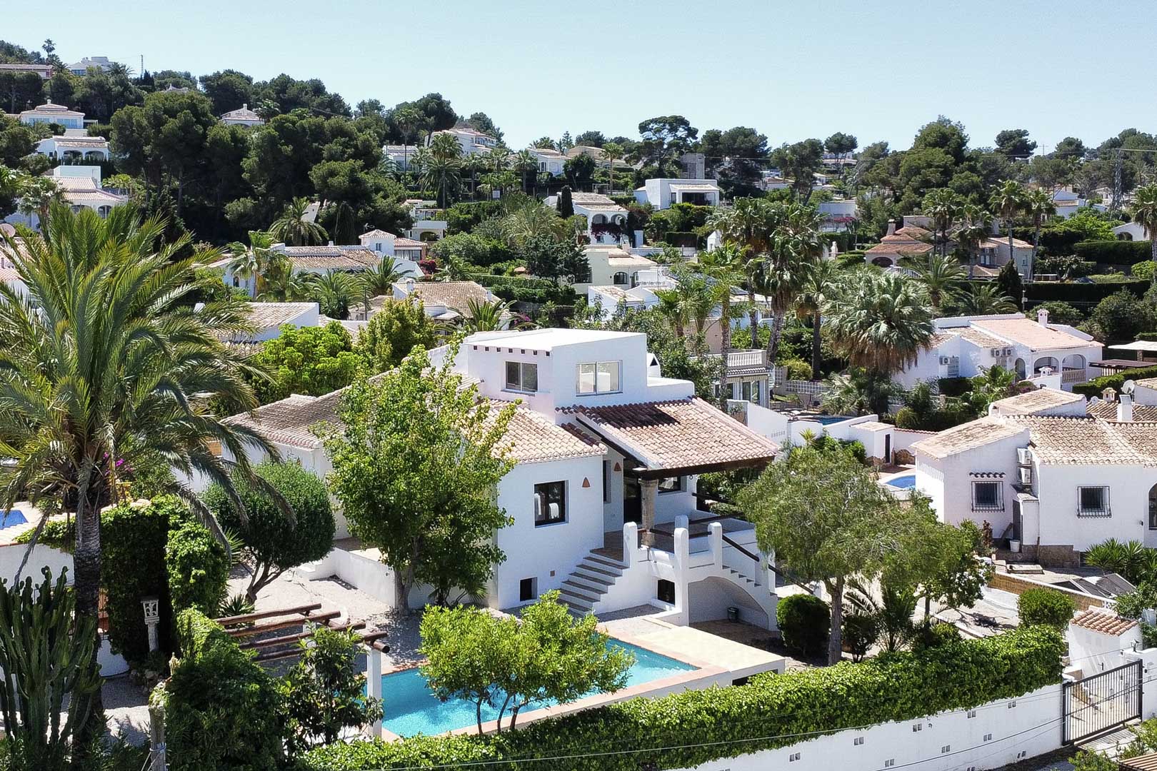 Elevated view of white villa with turquoise pool and tile roof, nestled in lush greenery in Jávea.