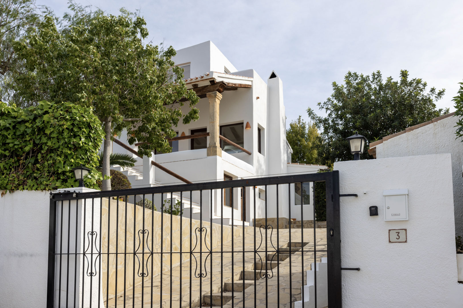 Exterior view of the villa featuring a wrought iron gate and white facade. Stone details and lush vegetation.