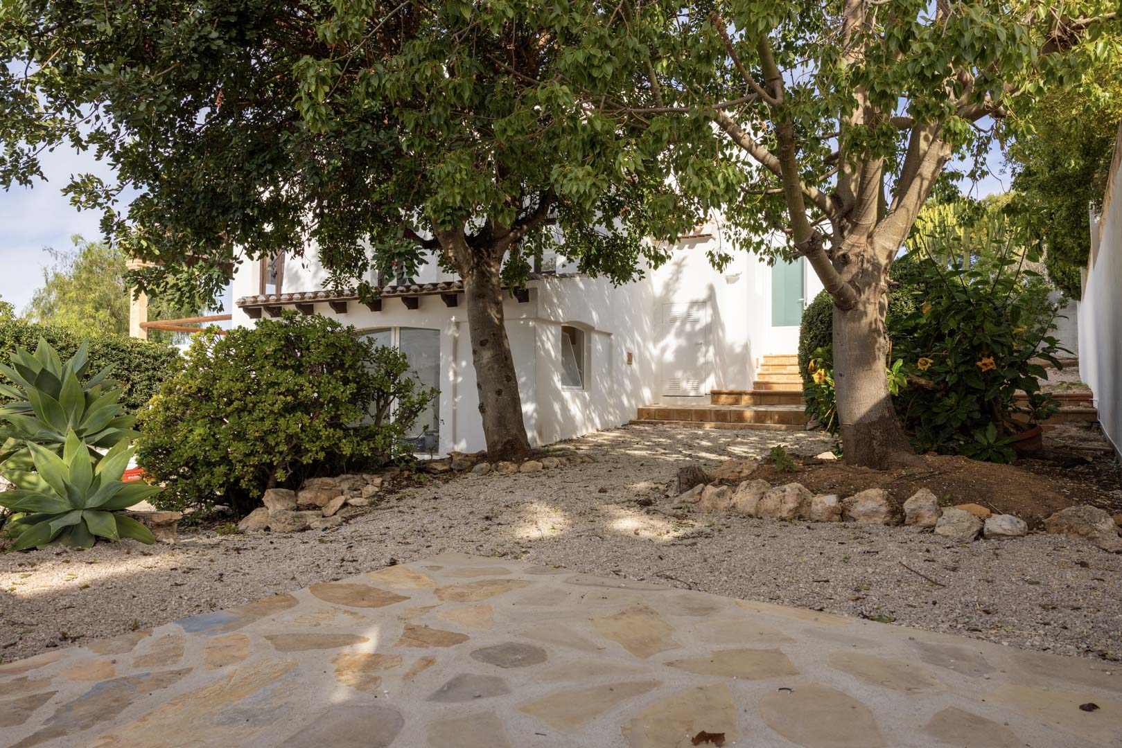 Exterior view of the villa with a gravel garden and access to the entrance. Mature greenery and white facade.