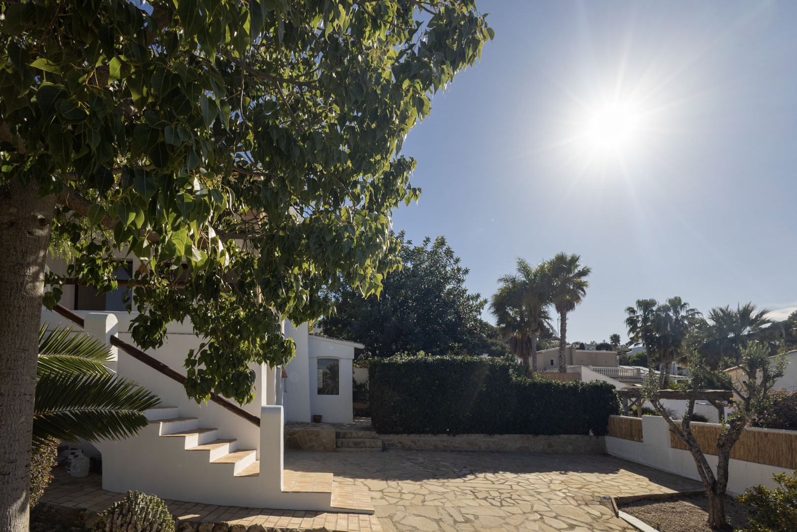 Villa exterior with stone patio, white stairs, and lush greenery under the sun.
