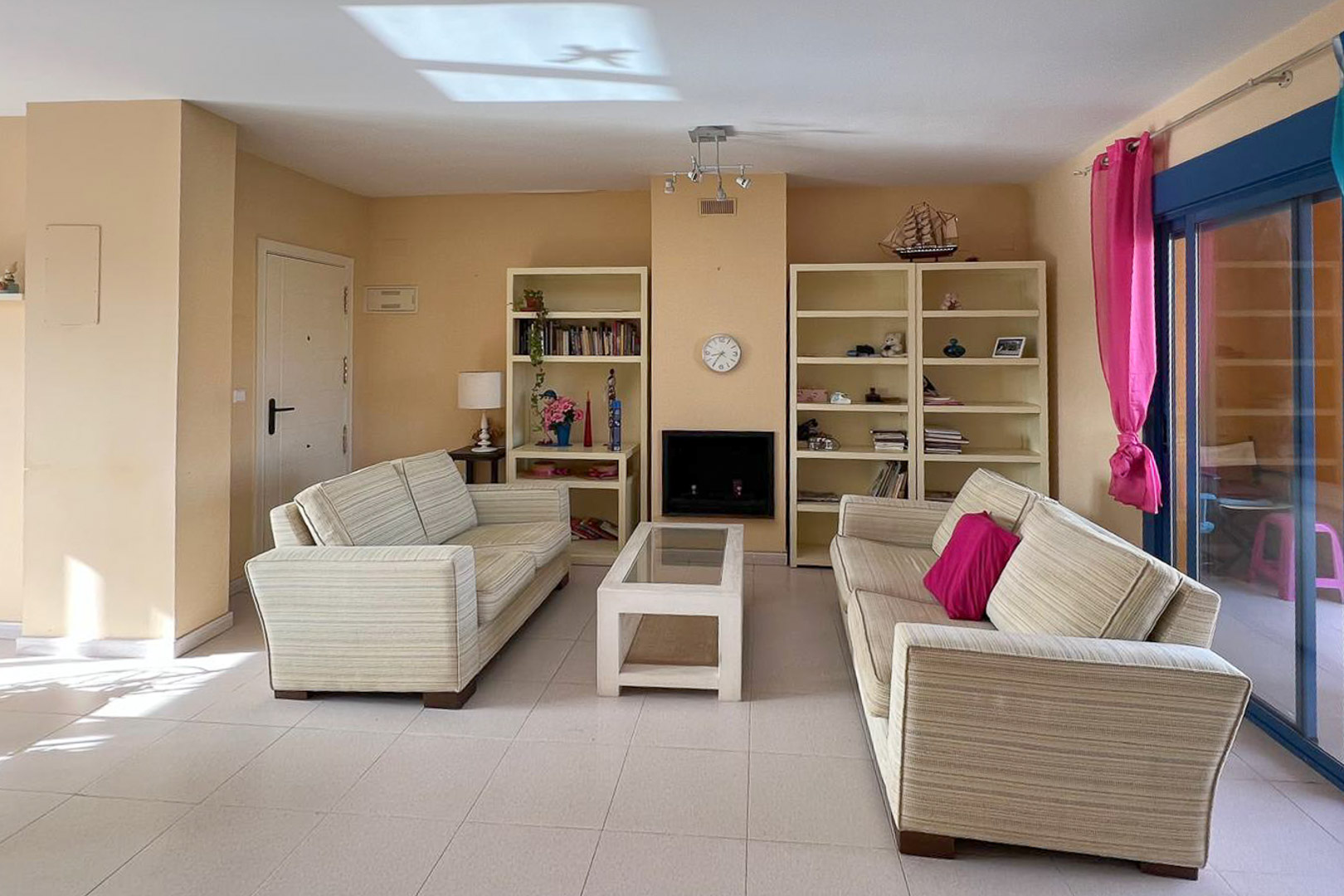Bright living room featuring striped sofas, white shelving units, and a decorative fireplace. Light tile flooring.