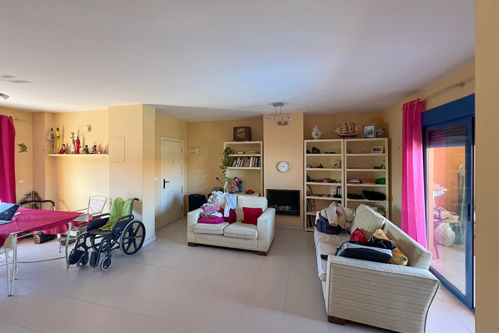 Living-dining area with shelving, upholstered sofas, and terrace access with pink curtains.
