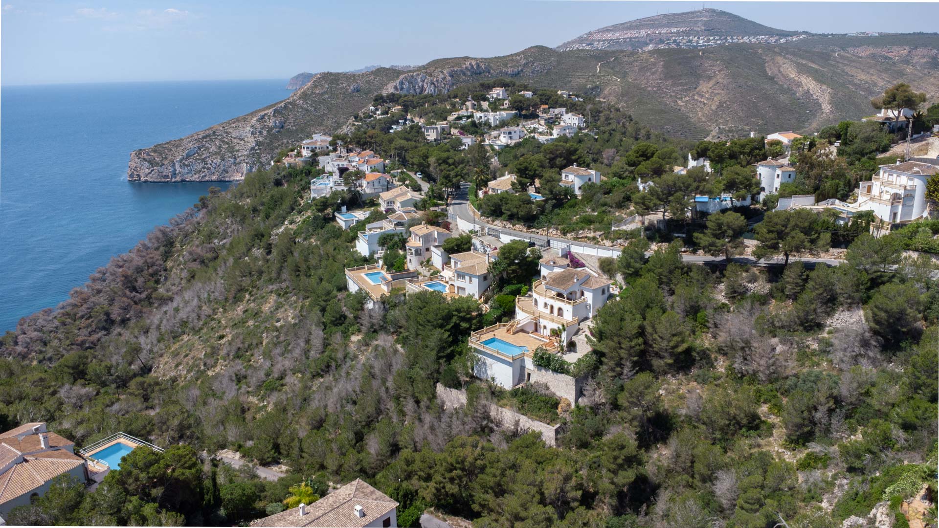 Aerial view of the Jávea coastline featuring residential villas nestled in a wooded hillside overlooking the Mediterranean Sea and rugged cliffs.