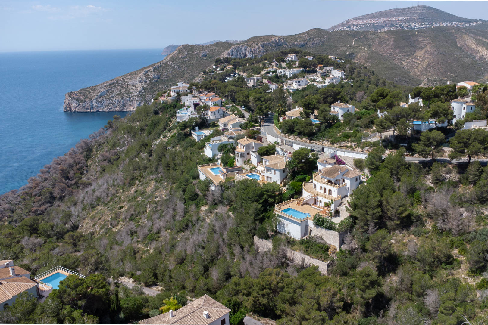 Aerial view of the Costa Nova urbanization in Jávea, featuring white villas with private pools nestled in a pine forest overlooking the Mediterranean coast.